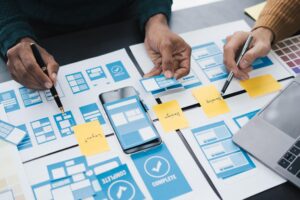 Team members planning a mobile app interface with wireframes, sticky notes, and smartphone mockups, illustrating how to create a social media platform through UI/UX design, user experience planning, and social networking app development.